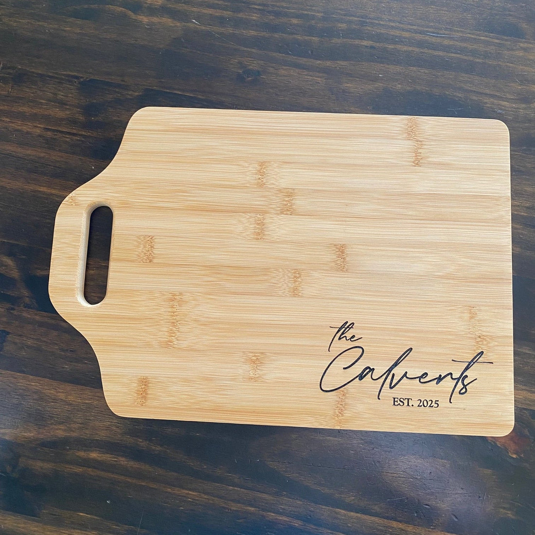 Bamboo handle Cutting board with family last name and est. date added underneath name. Cutting is displayed on a dark wood counter background. 