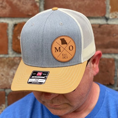 Leather Missouri Patch shown on a Richardson 112 trucker hat in heather grey with amber gold bill and birch mesh with a model wearing a blue shirt with a red brick background behind him