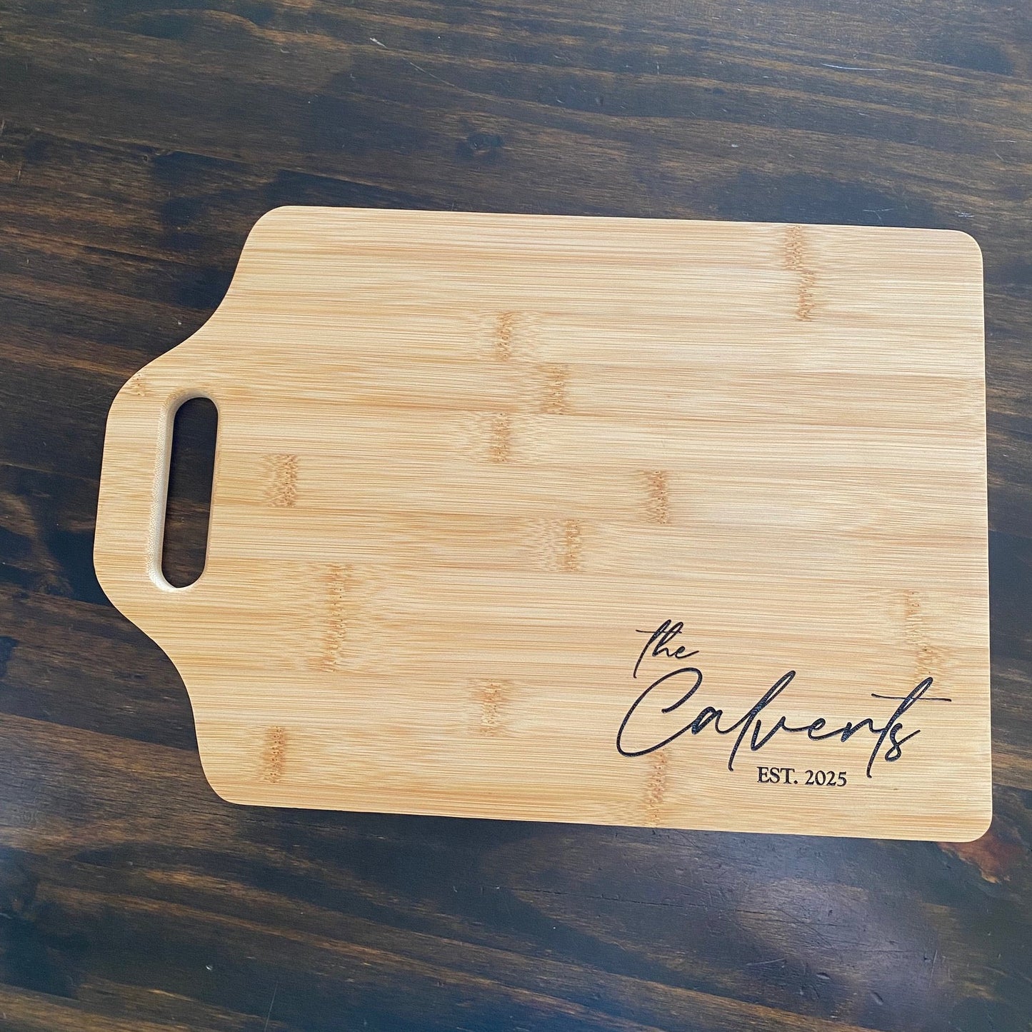 Wooden cutting board with 'the Calverts EST. 2023' engraving on a dark wooden surface
