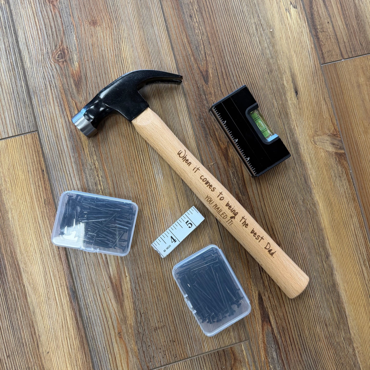 Hammer with engraved text on a wooden floor, surrounded by small containers and a level.