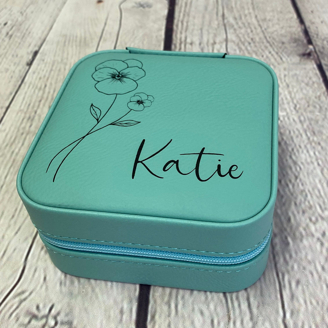 Teal jewelry box with 'Katie' and floral designs on a wooden surface
