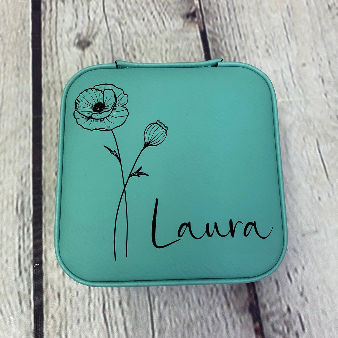 Teal square pouch with floral design and 'Laura' text on a wooden surface