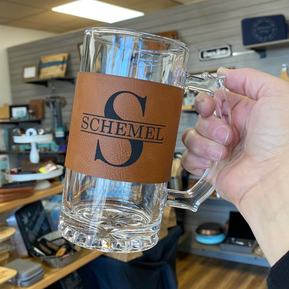 Clear glass mug with a brown leather tag featuring 'Schemel' customization held by a hand.
