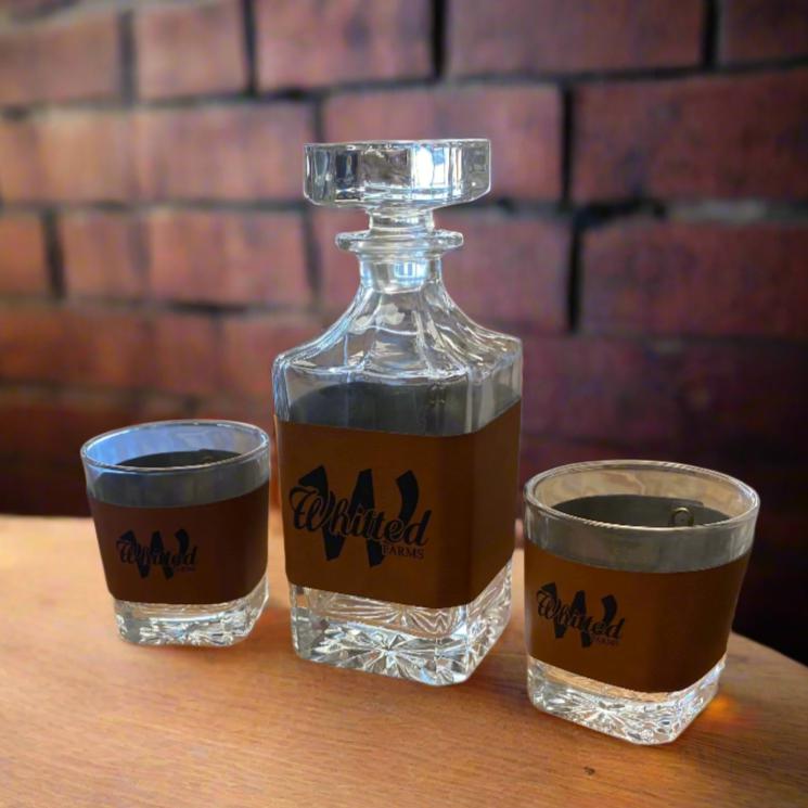 A leather-wrapped decanter with a glass stopper, alongside two bourbon glasses, all with a personalized logo on the decanter.