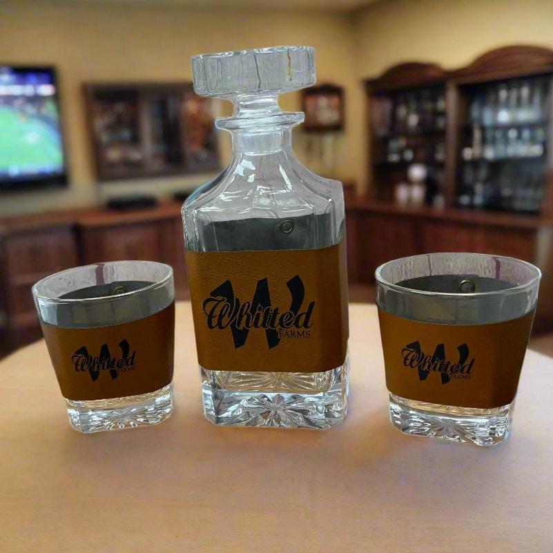 A leather-wrapped decanter with a glass stopper, alongside two bourbon glasses, all with a personalized logo on the decanter.