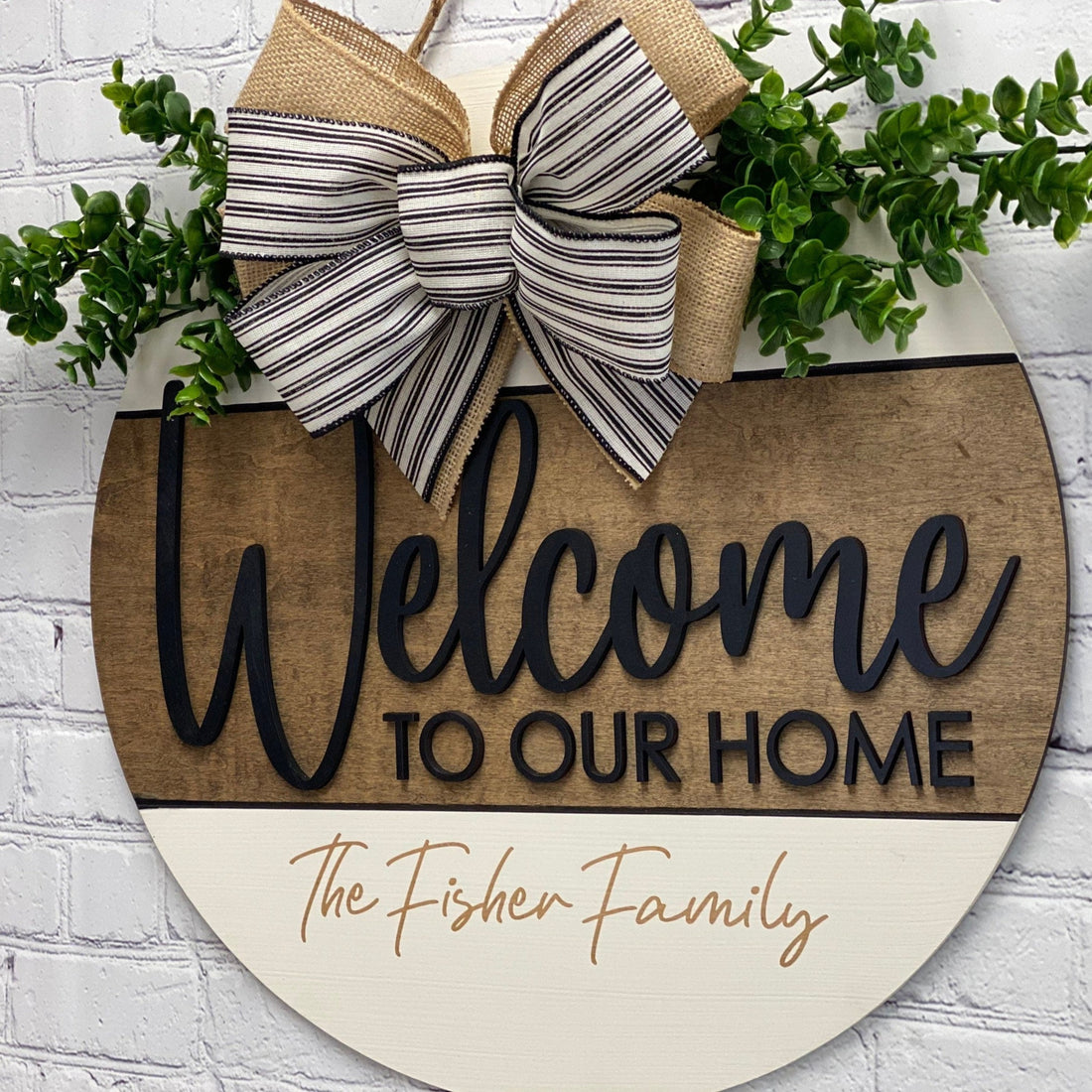 A round wooden door hanger with a bow and greenery on top, featuring the text 'Welcome to our Home' in a 3D effect, along with a small family name below the text.