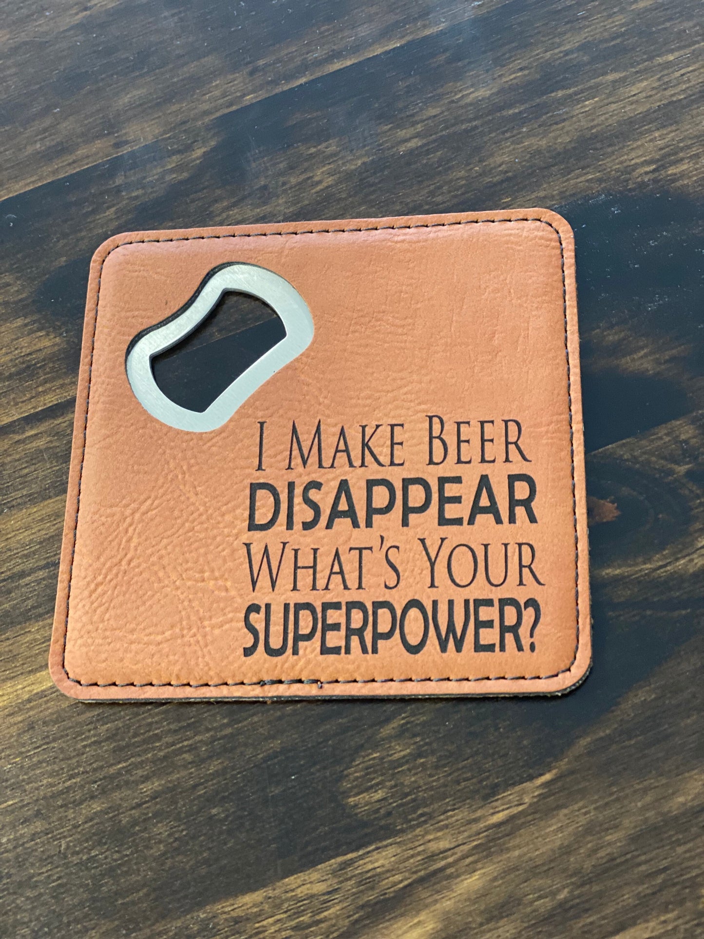 A brown leatherette coaster bottle opener with a stainless steel spike and the phrase 'I make beer disappear what's your superpower?' engraved on it.