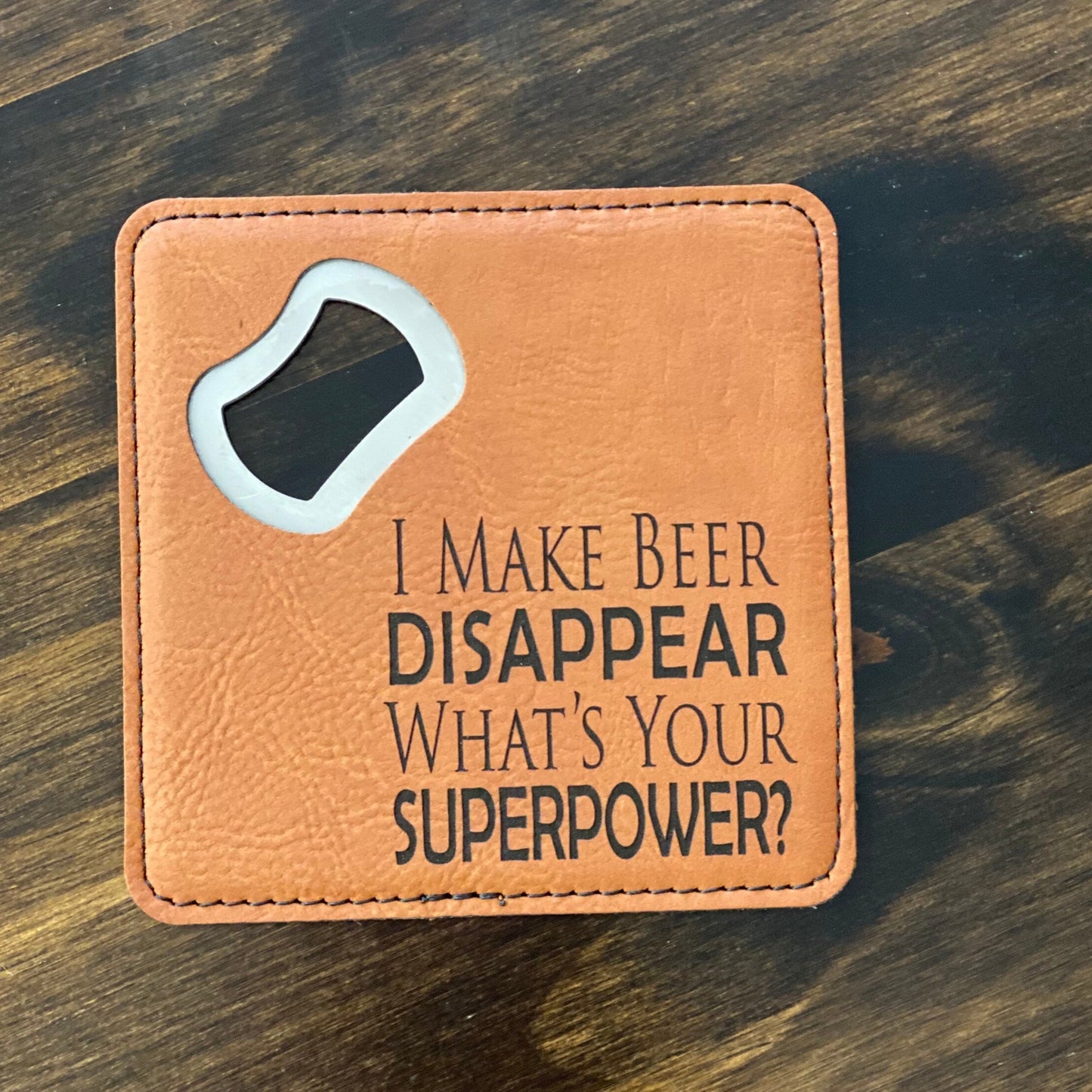 A brown leatherette coaster bottle opener with a stainless steel spike and the phrase 'I make beer disappear what's your superpower?' engraved on it.
