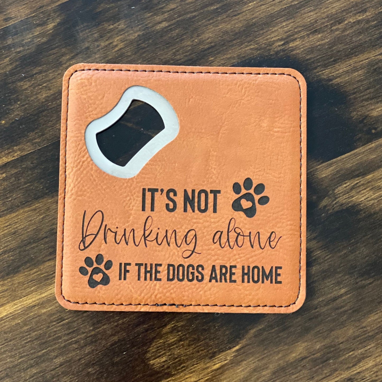 A brown leatherette coaster bottle opener with a stainless steel spike and the text 'It's Not Drinking Alone If Dogs Are Home' engraved on the front.