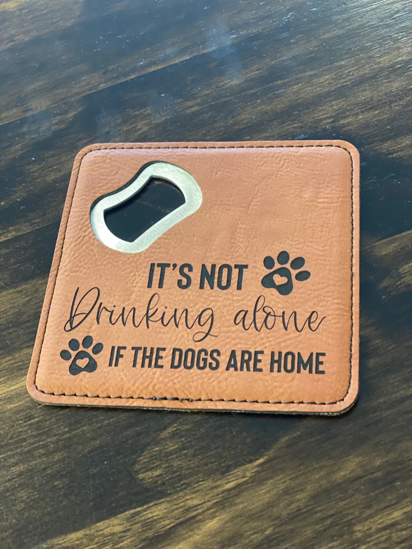 A brown leatherette coaster bottle opener with a stainless steel spike and the text 'It's Not Drinking Alone If Dogs Are Home' engraved on the front.