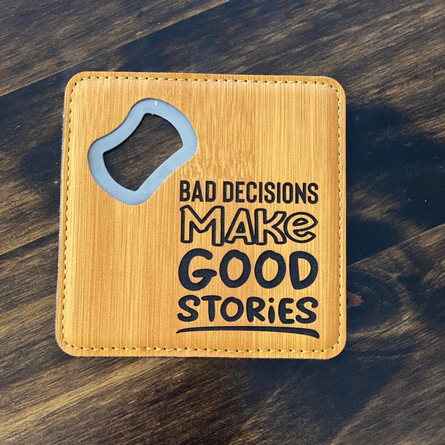 A brown leatherette coaster bottle opener with a stainless steel spike and the text 'Bad decisions make good stories' engraved on the front.