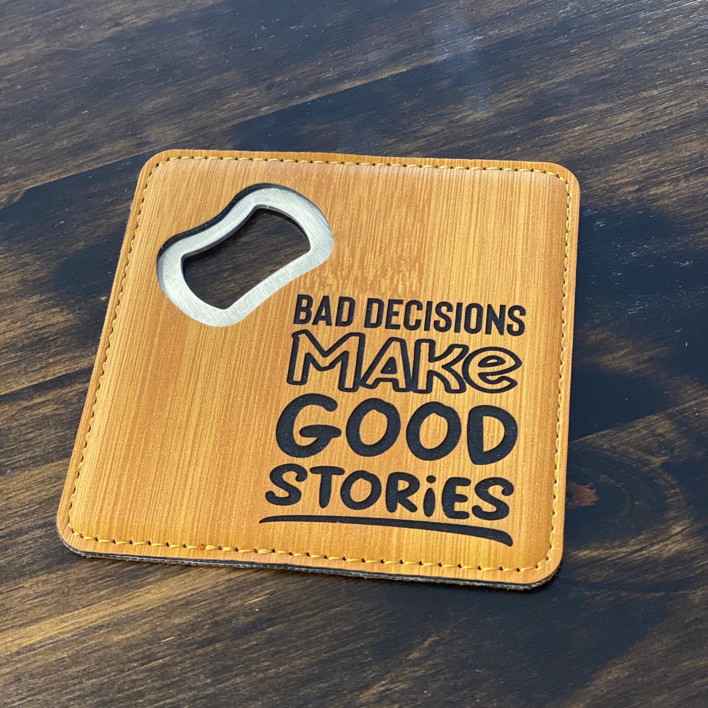 A brown leatherette coaster bottle opener with a stainless steel spike and the text 'Bad decisions make good stories' engraved on the front.